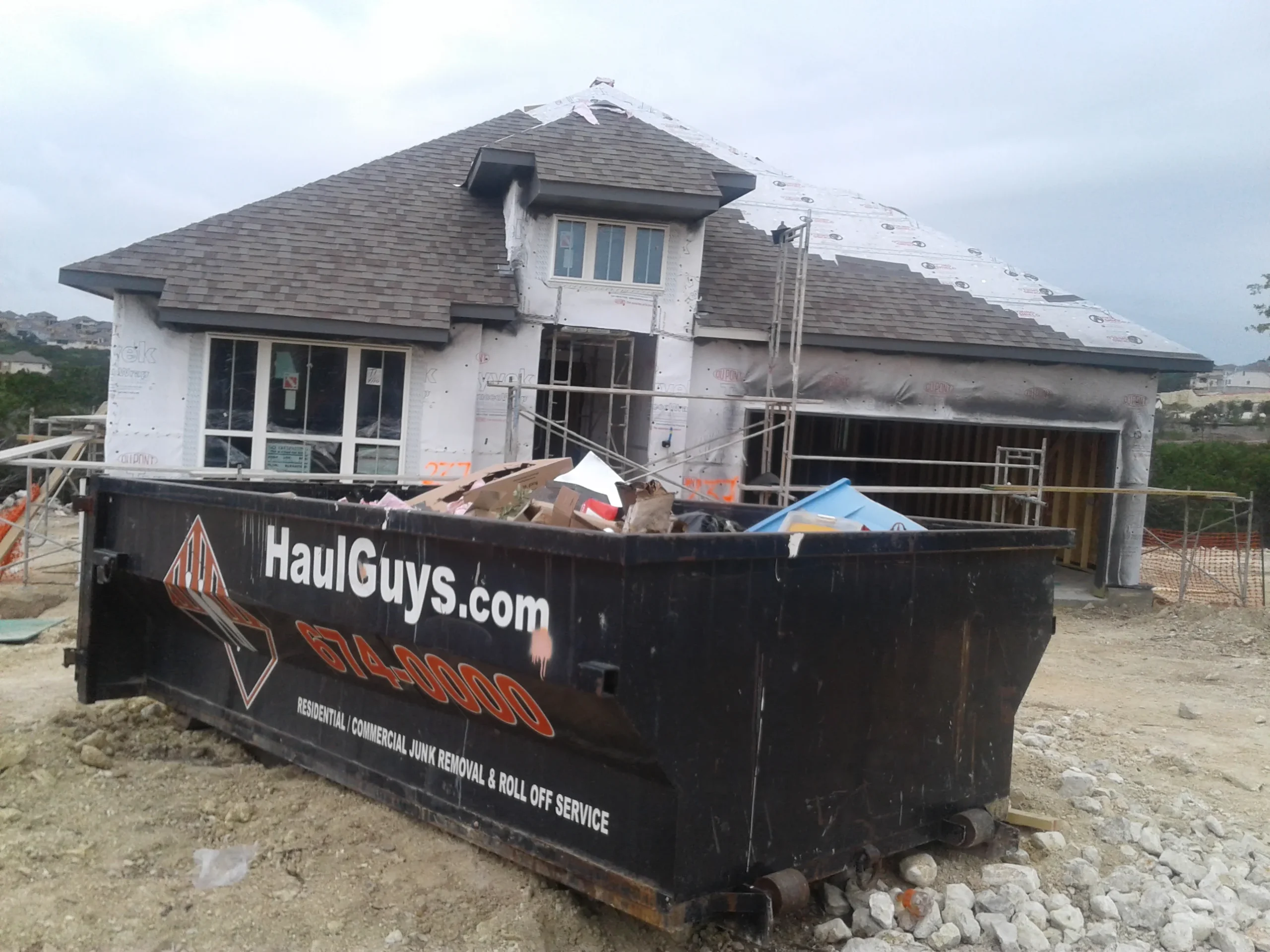 A Haul Guys dumpster filled with construction debris, parked in front of a house under construction with scaffolding and building materials on-site.