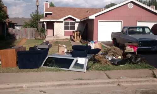 A residential property with various discarded items, including furniture, boxes, and debris, scattered on the front lawn and driveway, awaiting removal. A pickup truck is parked in the driveway beside the items.
