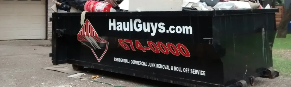 A Haul Guys trailer loaded with various junk materials and covered by a black tarp, featuring the company's logo and contact number, parked in a residential area surrounded by trees.